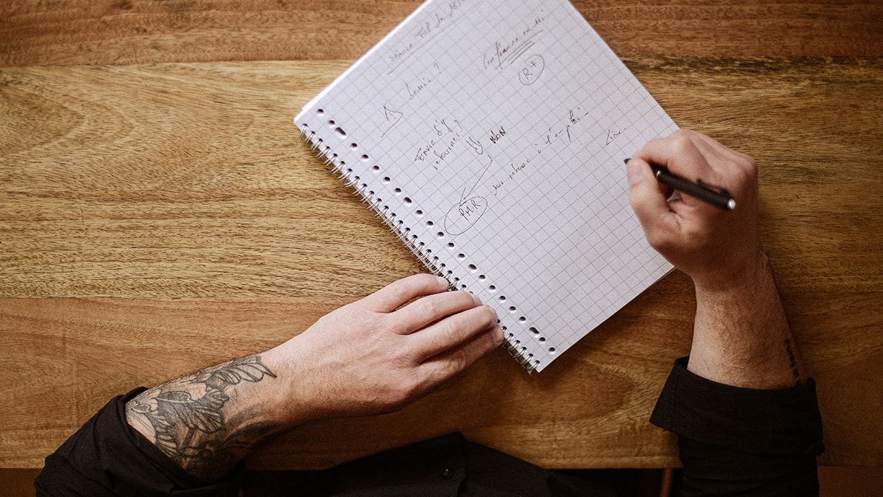 propulse Personne tatouée prenant des notes manuscrites sur un cahier quadrillé posé sur une table en bois